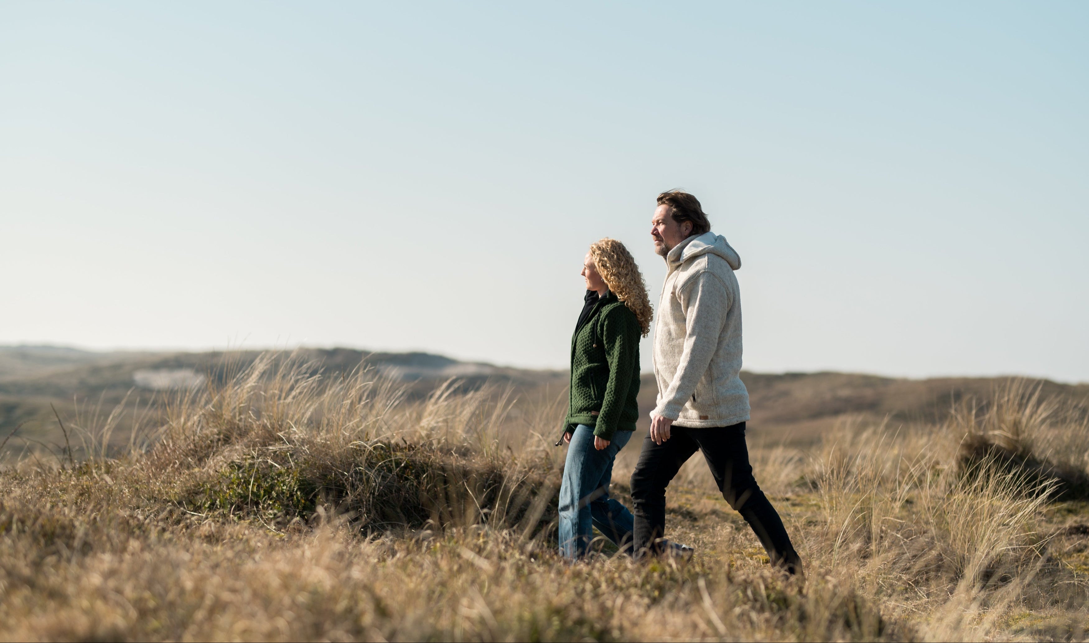 Een mannelijk- en vrouwelijk model wandelen samen door de duinen met een wollen Puur Nepal vest aan